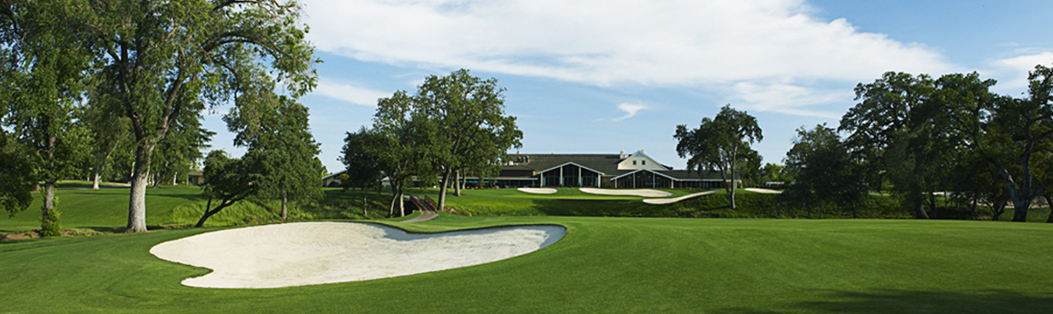 Golf course landscape with curved white sand bunkers, green fairway, trees, and a low clubhouse building under a partly cloudy sky