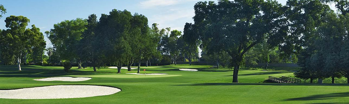 Lush green golf course fairway with sand bunkers, trees, and a flag, under blue sky